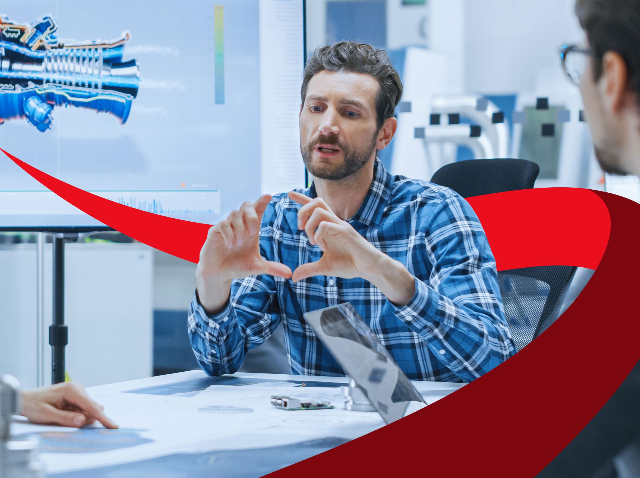 Technical professional explaining something to others at a conference table with a computer screen behind him.